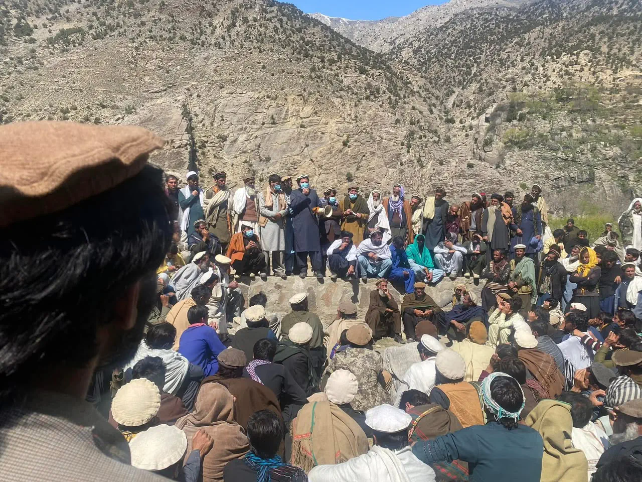A large jirga in Mandol: men of the tribe seated on the ground and standing along a stone wall below a rocky mountain, a speaker with a loudhailer in the middle