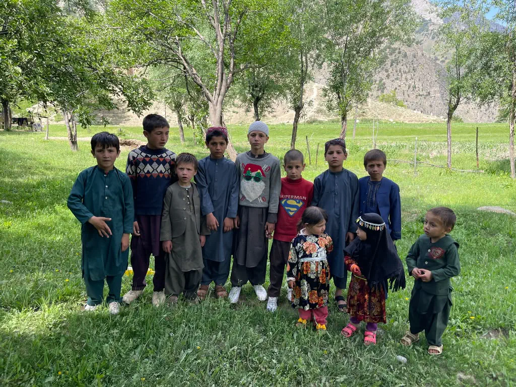 A group of children in Kantiwa standing together on the grass under trees, two small girls in bright embroidered dresses in front