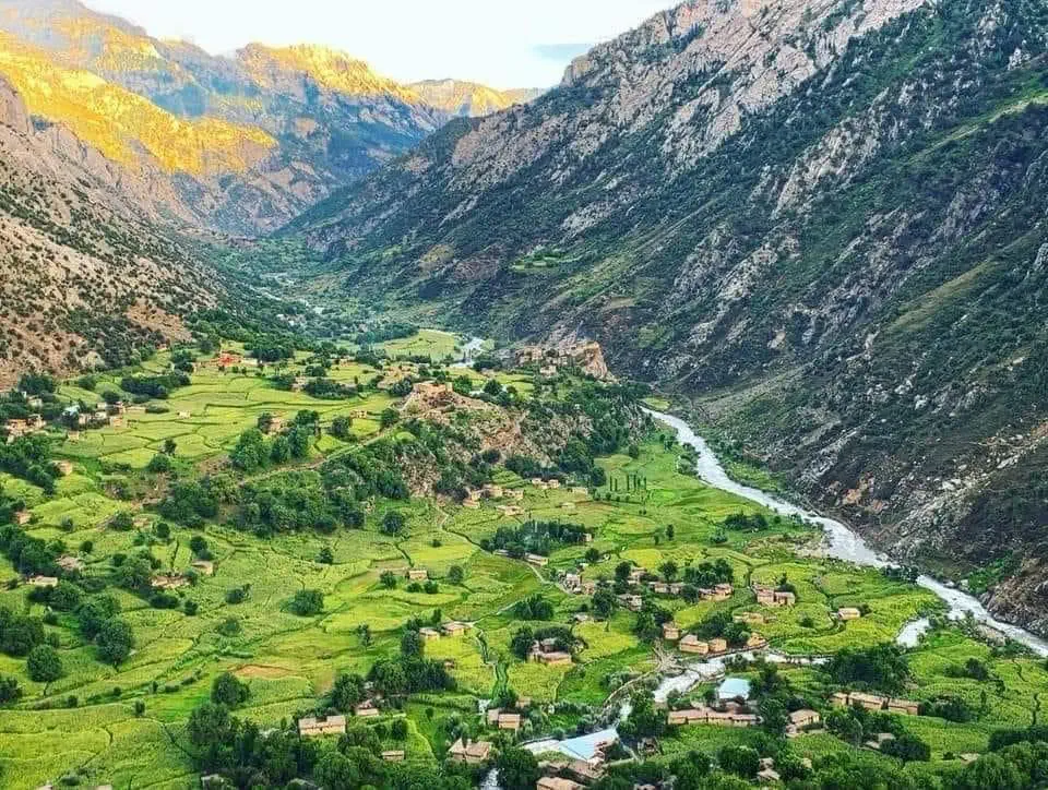 The Kantiwa valley in Nuristan from above: green fields and scattered houses along a winding river, steep rocky mountains on both sides, the peaks lit gold