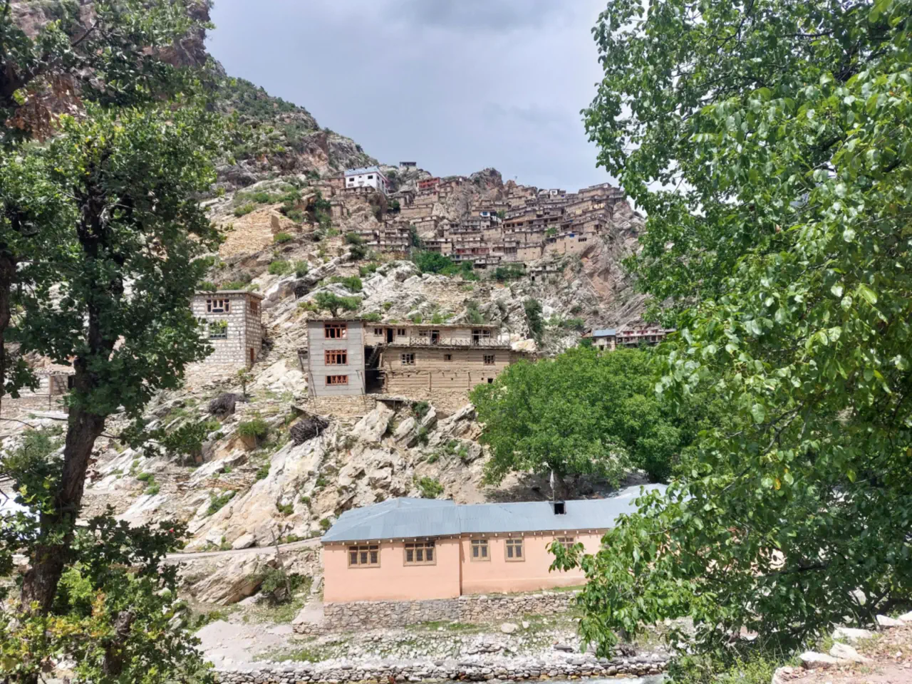 Stone and timber houses stacked up a rocky hillside in Waygal, a pink-walled building with a blue roof below among trees