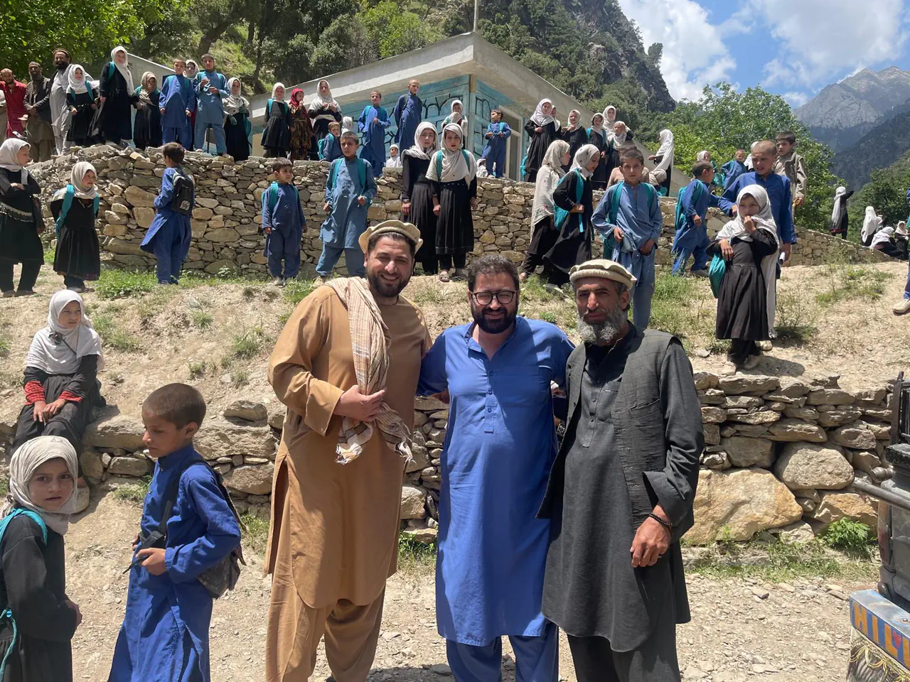 Zabiullah Amiri with two companions in front of a village school in the Yinshaw valley of Waygal district, dozens of pupils in blue and white on the stone terraces behind