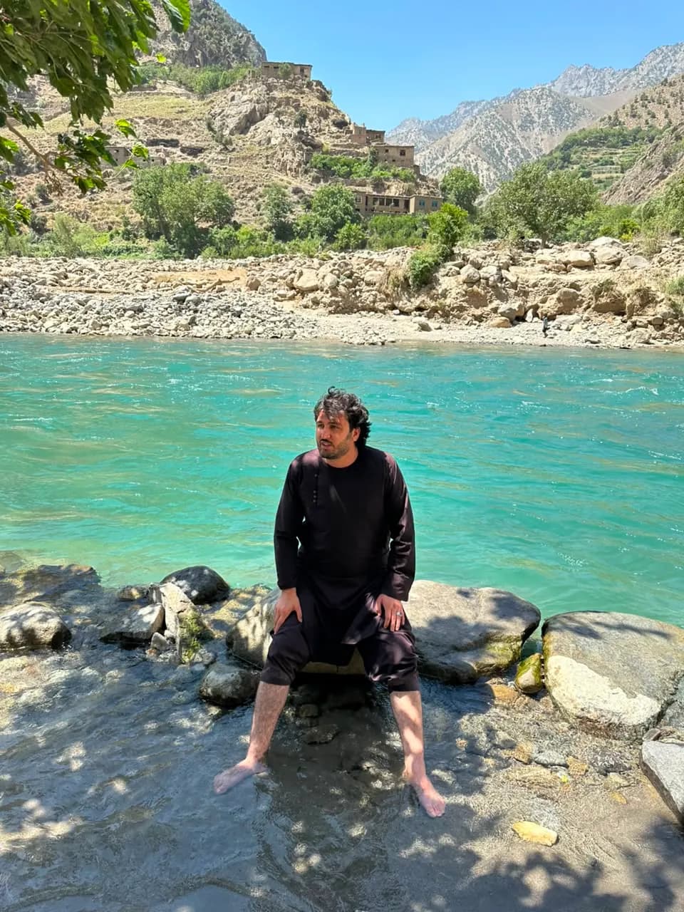 Abdulrahman Chonish sitting on rocks with his feet in a turquoise river, a hillside village behind