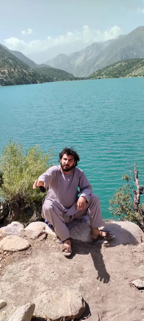 Abdulrahman Chonish crouched on rocks beside a turquoise lake ringed by mountains
