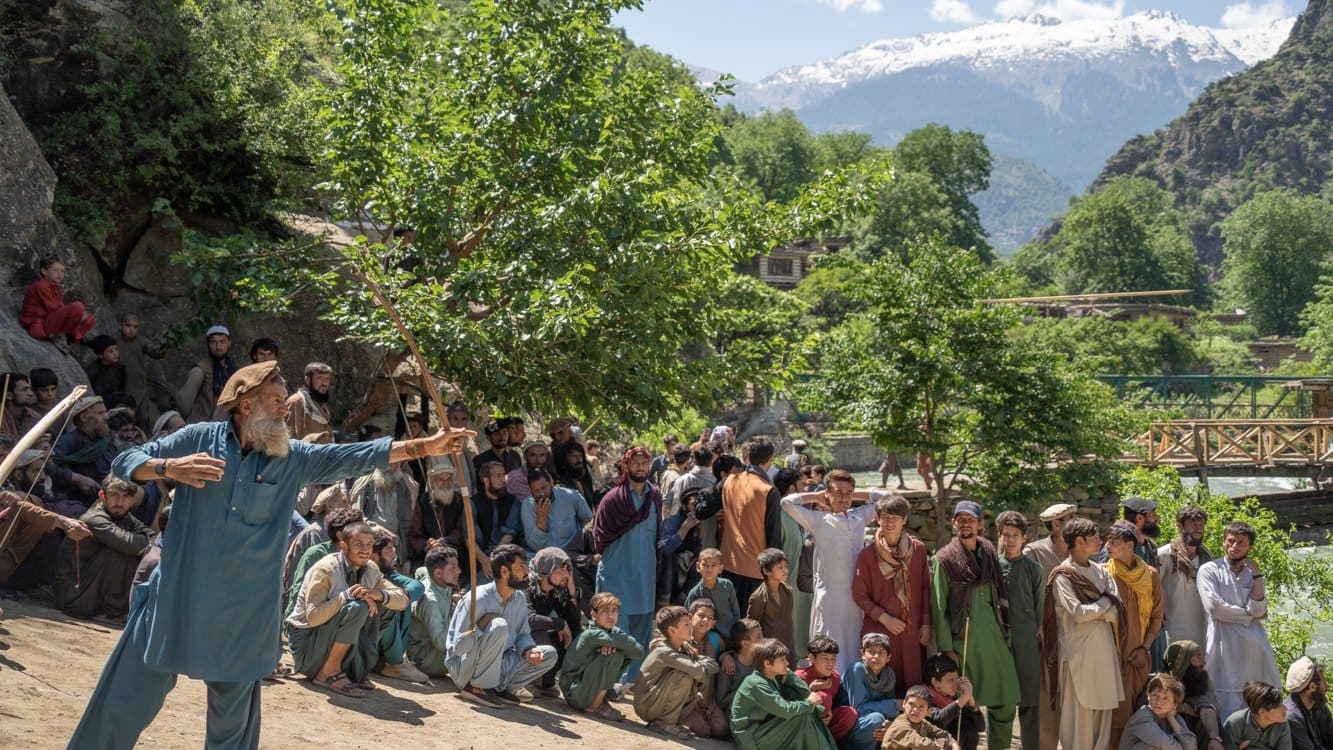 An elder loosing an arrow from a long wooden bow before a crowd by the river