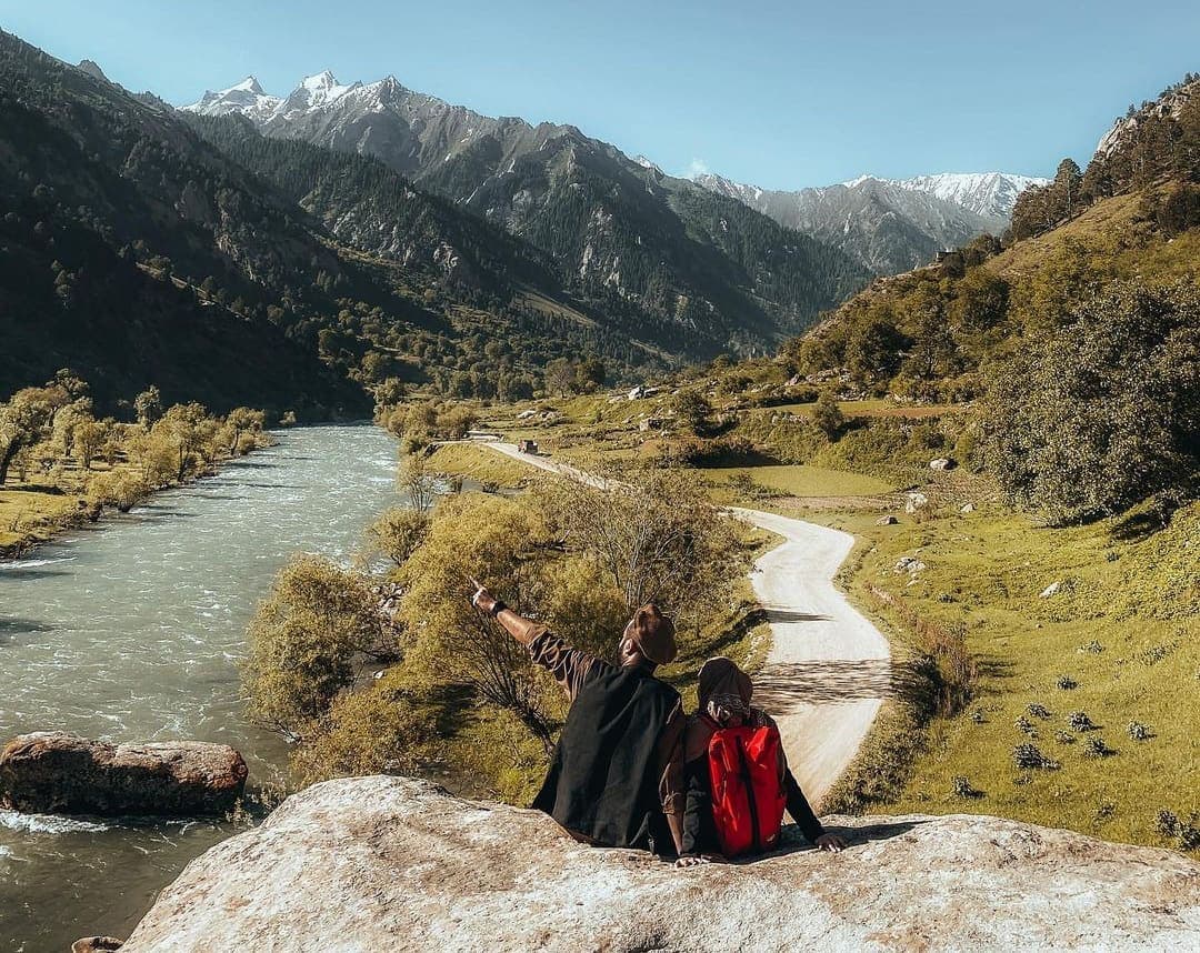Two travellers on a boulder above a wide river valley, snow peaks beyond