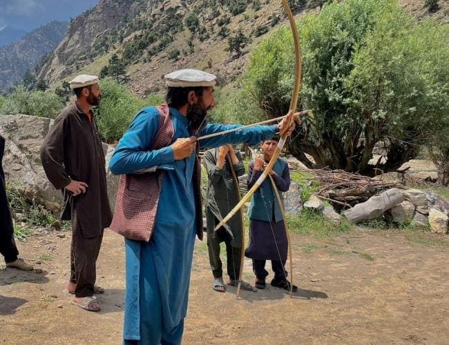 A man in a blue kameez drawing a long wooden bow as others watch