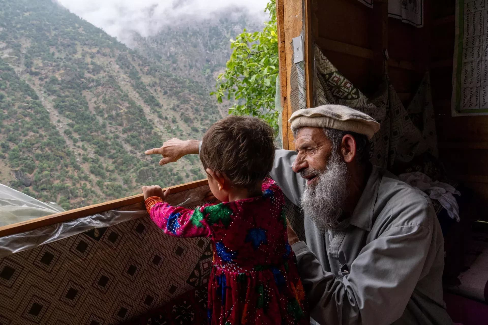 A Nuristani elder in a pakol and a child in embroidered dress looking out over the mountains from their home