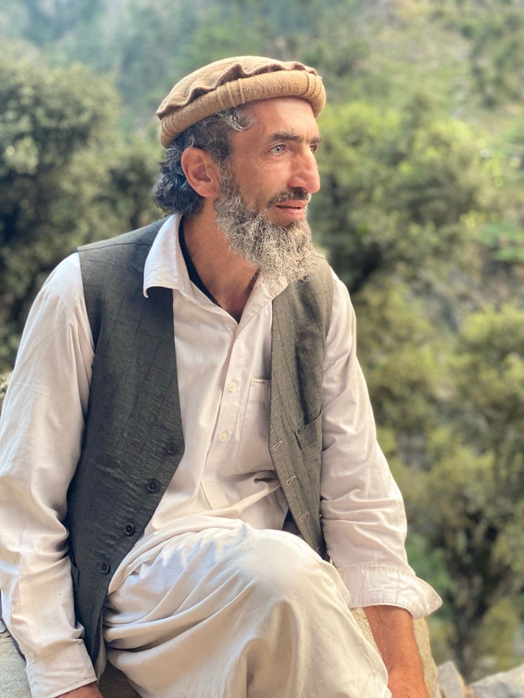 A grey-bearded man in a tan pakol and grey waistcoat, forest behind him