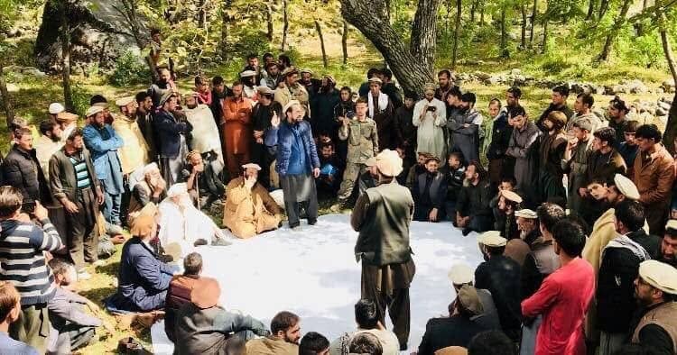 A large circle of men seated and standing at an outdoor village council