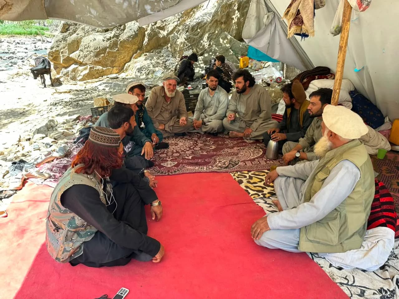 Men seated in a circle on rugs under a canvas shelter, talking over tea