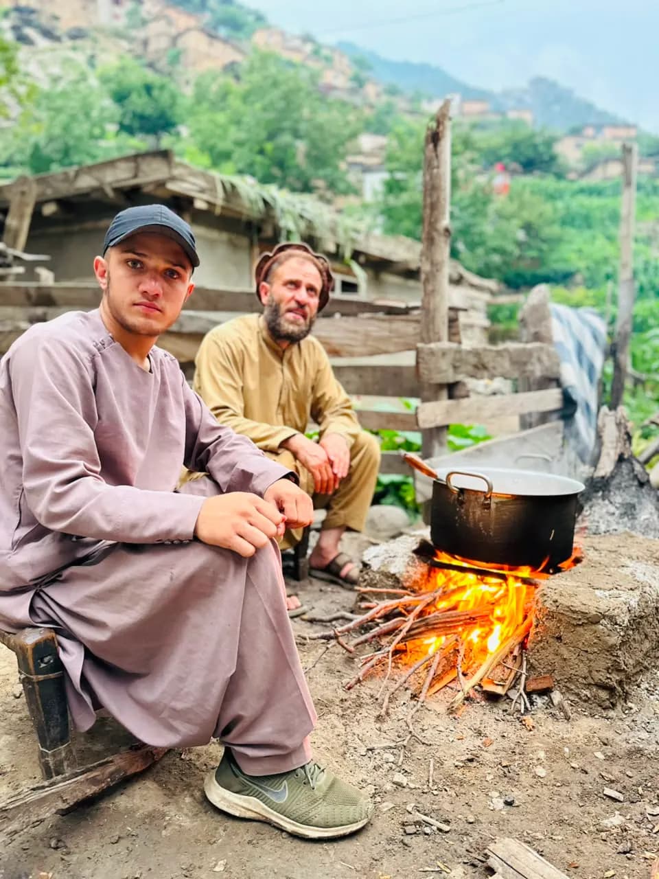 Two men cooking over an open wood fire in Waygal, village slopes behind