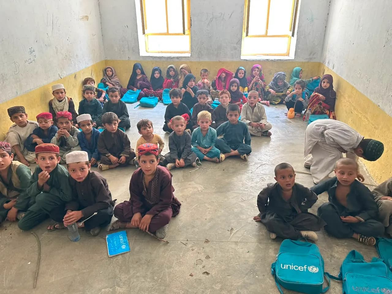 Children seated on the floor of a village classroom with school bags
