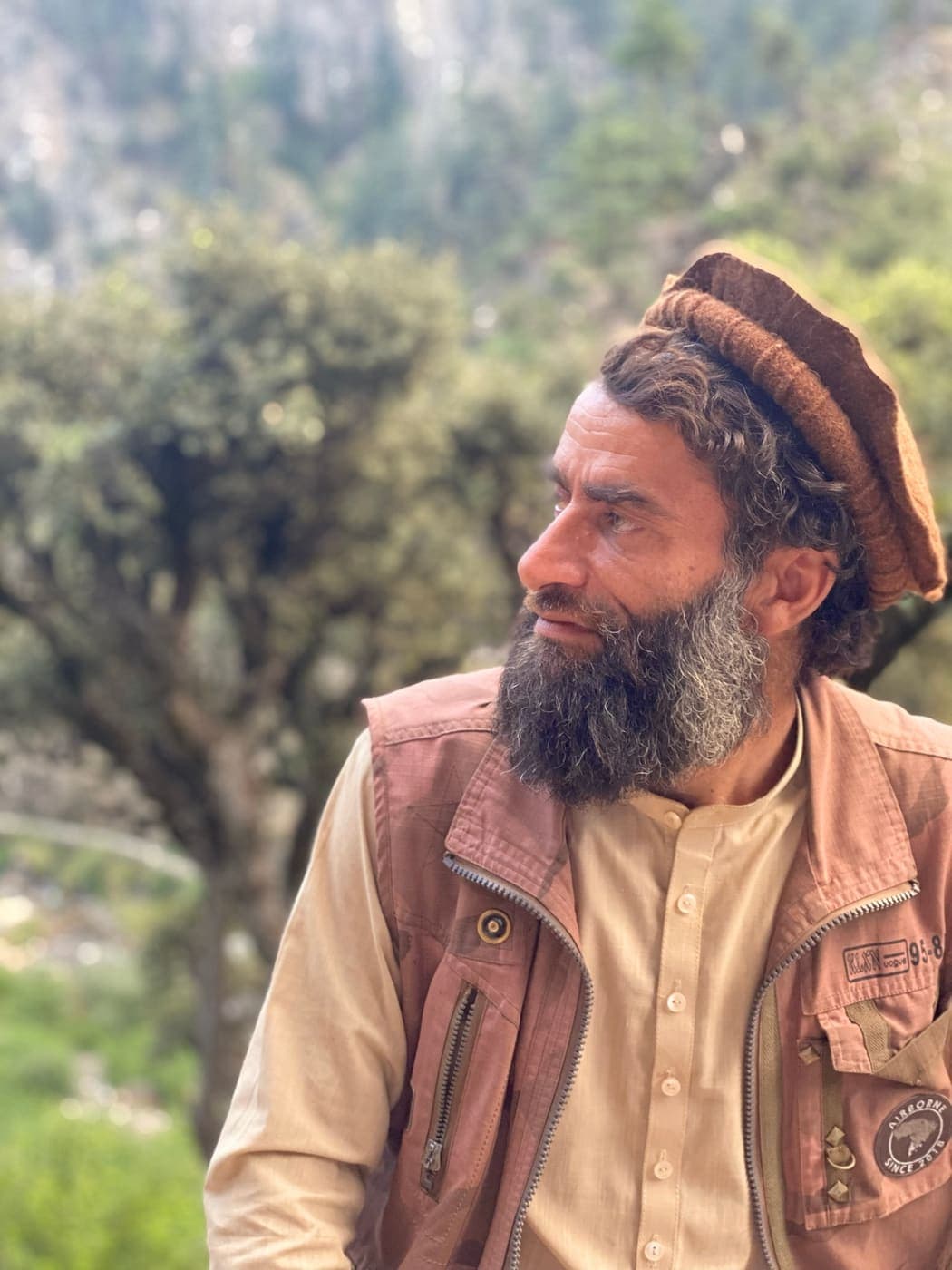 A bearded Nuristani man in a pakol and utility vest in the mountains
